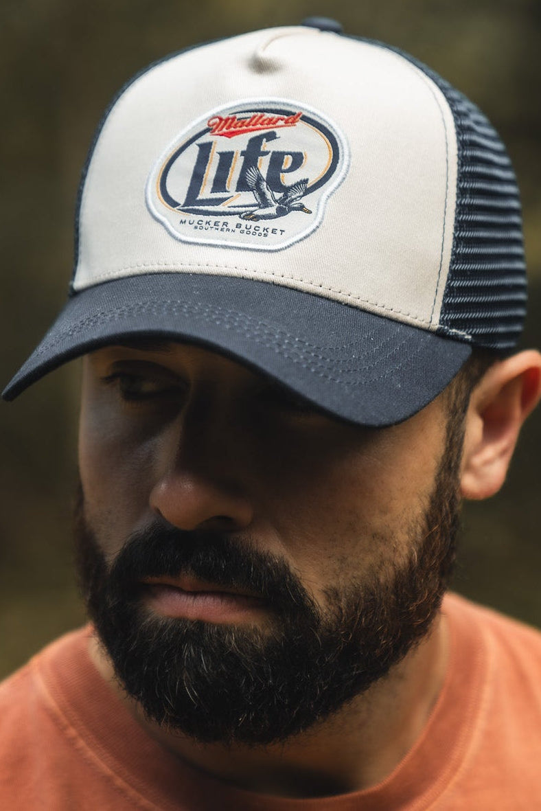 A bearded man wearing a navy and white Mucker Bucket Mallard Life Trucker Bucket hat and an orange shirt, looking slightly down and to the side outdoors with a blurred natural background.