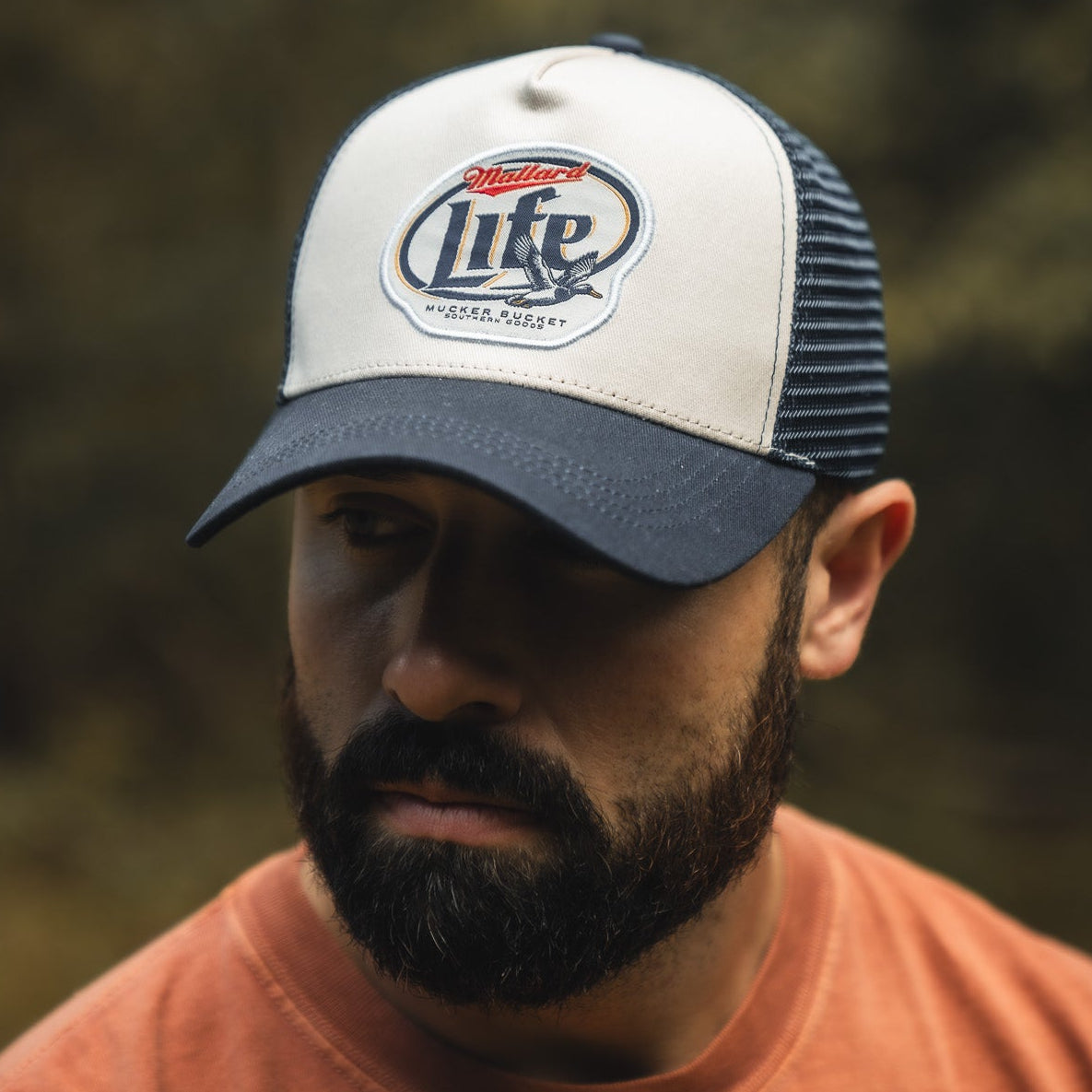A bearded man wearing the Mallard Life Trucker Bucket by My Store, a navy and white snapback with patch embroidery, looks downward outdoors in an orange shirt against a blurred natural background.