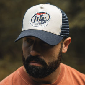 A bearded man wearing the Mallard Life Trucker Bucket by My Store, a navy and white snapback with patch embroidery, looks downward outdoors in an orange shirt against a blurred natural background.