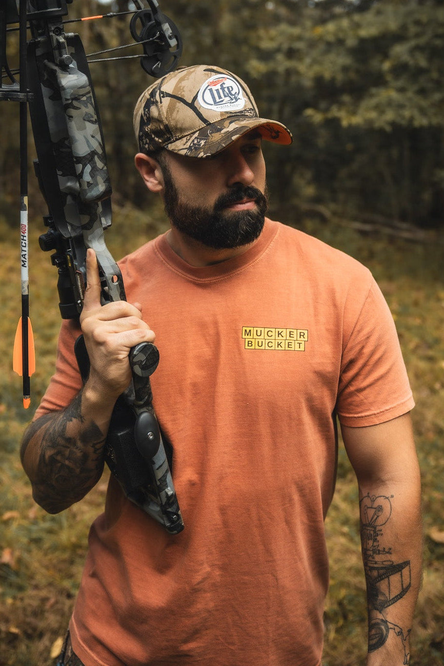 A bearded man in an orange Mucker Bucket t-shirt and a Mucker Bucket Mallard Life Deadwood Camo Bucket hat stands outdoors with a compound bow over his shoulder, surrounded by trees and grass.