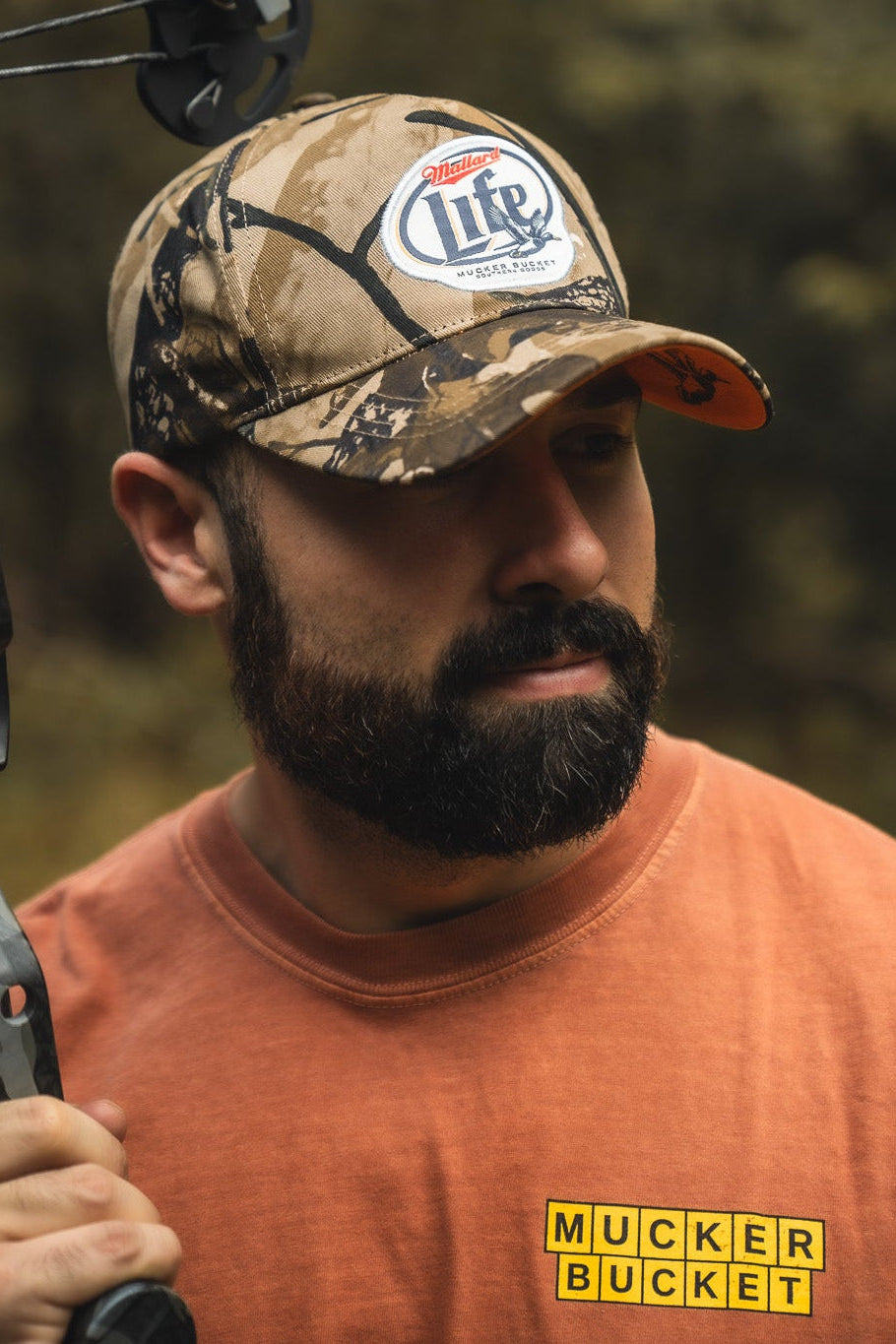 A bearded man in a Mallard Life Deadwood Camo Bucket by Mucker Bucket and an orange shirt holds a camo crossbow outdoors, glancing to the side with a forest backdrop.