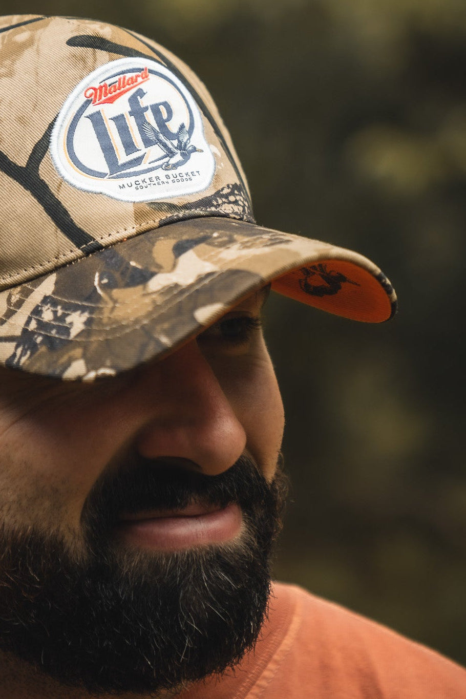 A man with a dark beard wears a Mucker Bucket Mallard Life Deadwood Camo Bucket hat. He’s in an orange shirt outdoors, with a blurred natural background that hints at mallard life.