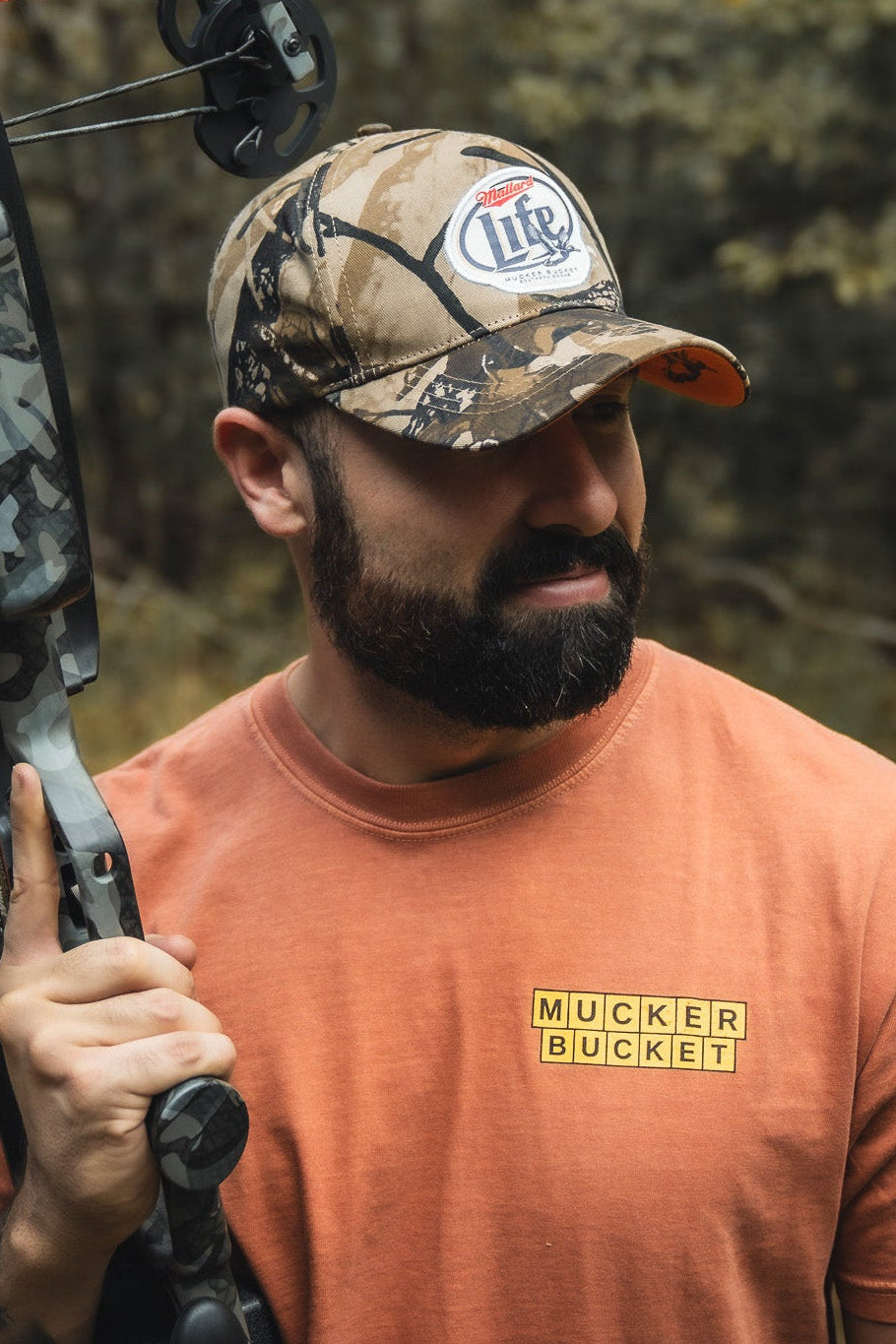 A bearded man in an orange Mucker Bucket shirt and a Mallard Life Deadwood Camo Bucket hat holds a camo-patterned crossbow over his shoulder while standing outdoors in a wooded area.