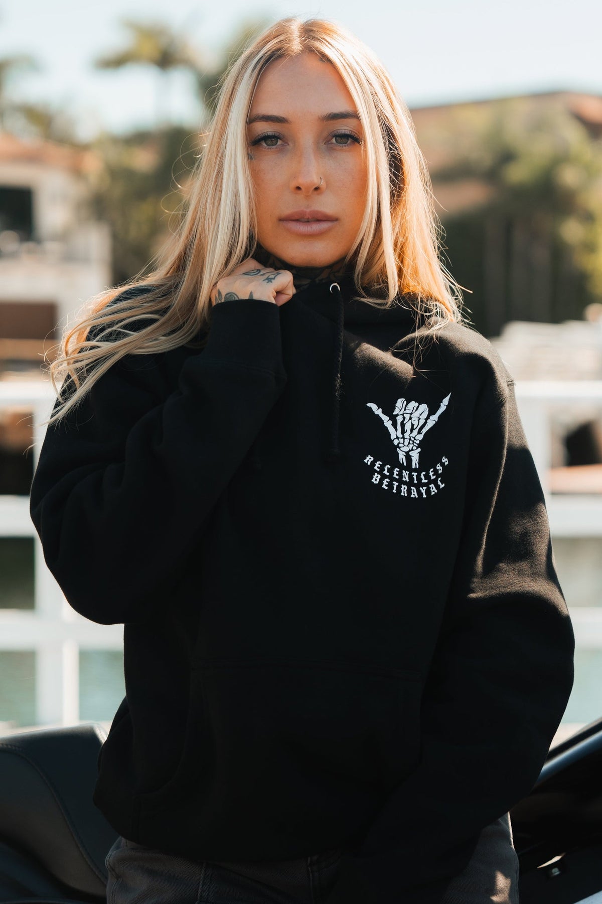 A woman with long blonde hair stands outdoors in a black Sink or Swim Premium Hoodie by Relentless Betrayal, featuring a skeletal hand graphic and resilience-themed text. She grips the collar with her right hand as the sunny, softly blurred background reflects her perseverance.