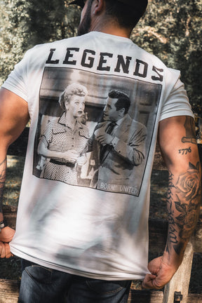 A man in a brown cap stands outdoors, showing the back of his white "Legends - Honey I'm Home!" t-shirt by Born United Private Label, featuring "LEGENDS" above a vintage black-and-white image, as he displays tattoos and holds honey pet treats.