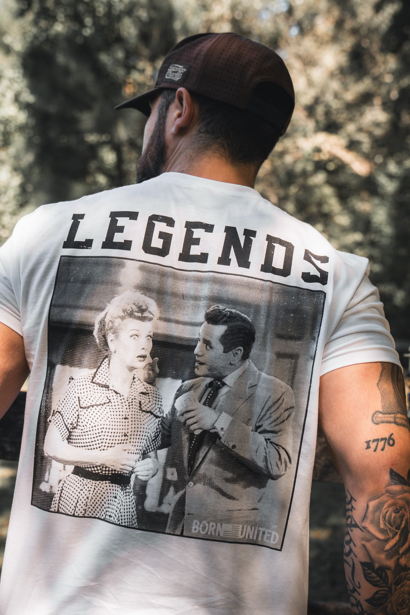 A man wears the "Legends - Honey I’m Home!" white t-shirt by Born United Private Label, featuring LEGENDS above a vintage couple. With tattoos, a brown cap, and pet treats in hand, he brings sweet charm to his casual style.