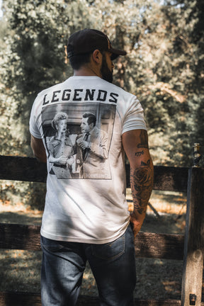 A man in a cap and white Legends - Honey I'm Home! t-shirt by Born United Private Label stands by a wooden fence outdoors, enjoying the sunlight. The shirt is perfect for grabbing pet treats or Honey on the go.
