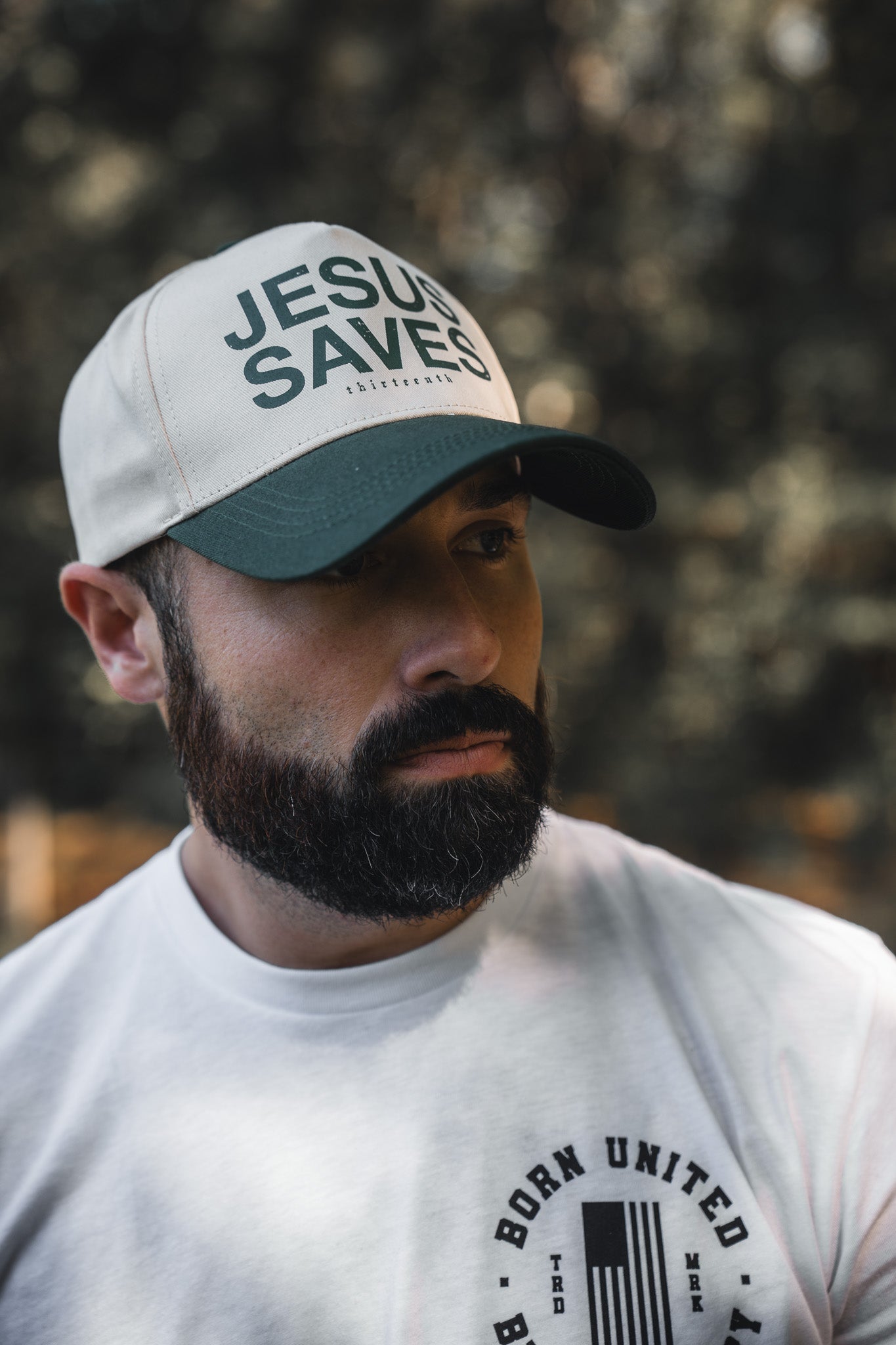 A bearded man wears a 13th Jesus Saves Ivory Snapback and a white tee with a circular BORN UNITED logo, standing outdoors against a blurred natural background that reflects the message of John 3:16.