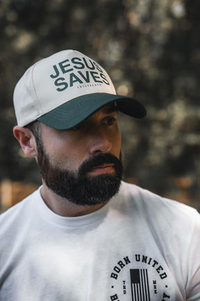 A bearded man wears a 13th Jesus Saves Ivory Snapback and a white tee with a circular BORN UNITED logo, standing outdoors against a blurred natural background that reflects the message of John 3:16.