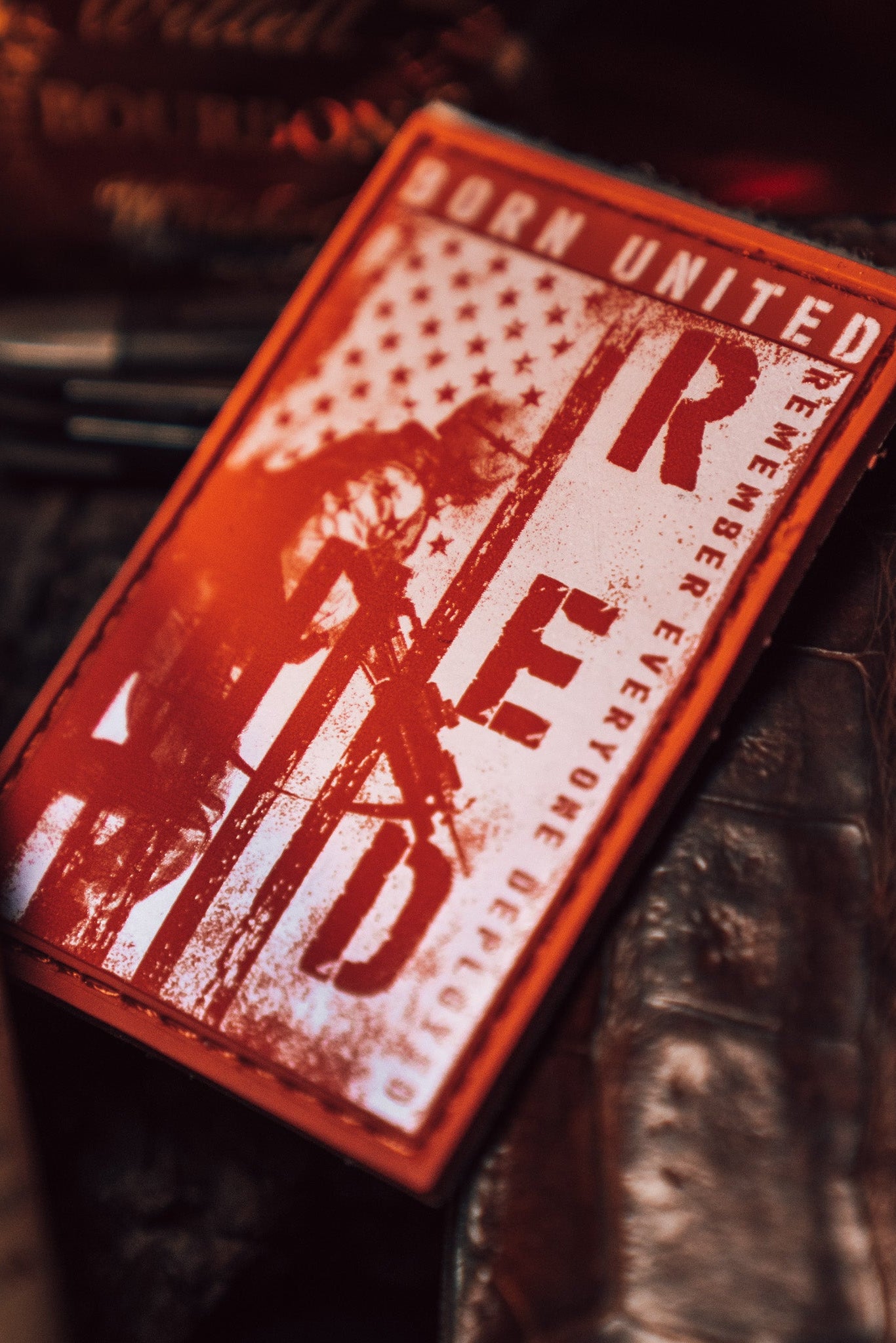 The R.E.D. Friday PVC Patch by Born United Private Label features a striking red and black design with R.E.D and Remember Everyone Deployed text, promoting military support. It includes a stylized American flag and soldier figure, surrounded by dark, blurred objects.