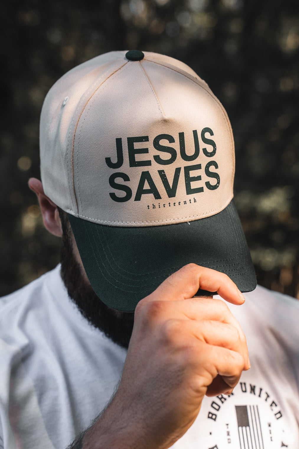 A person adjusts the brim of the 13th Jesus Saves Ivory Snapback outdoors, their face partially hidden by the cap—embroidered with "JESUS SAVES" and referencing John 3:16—and their hand.
