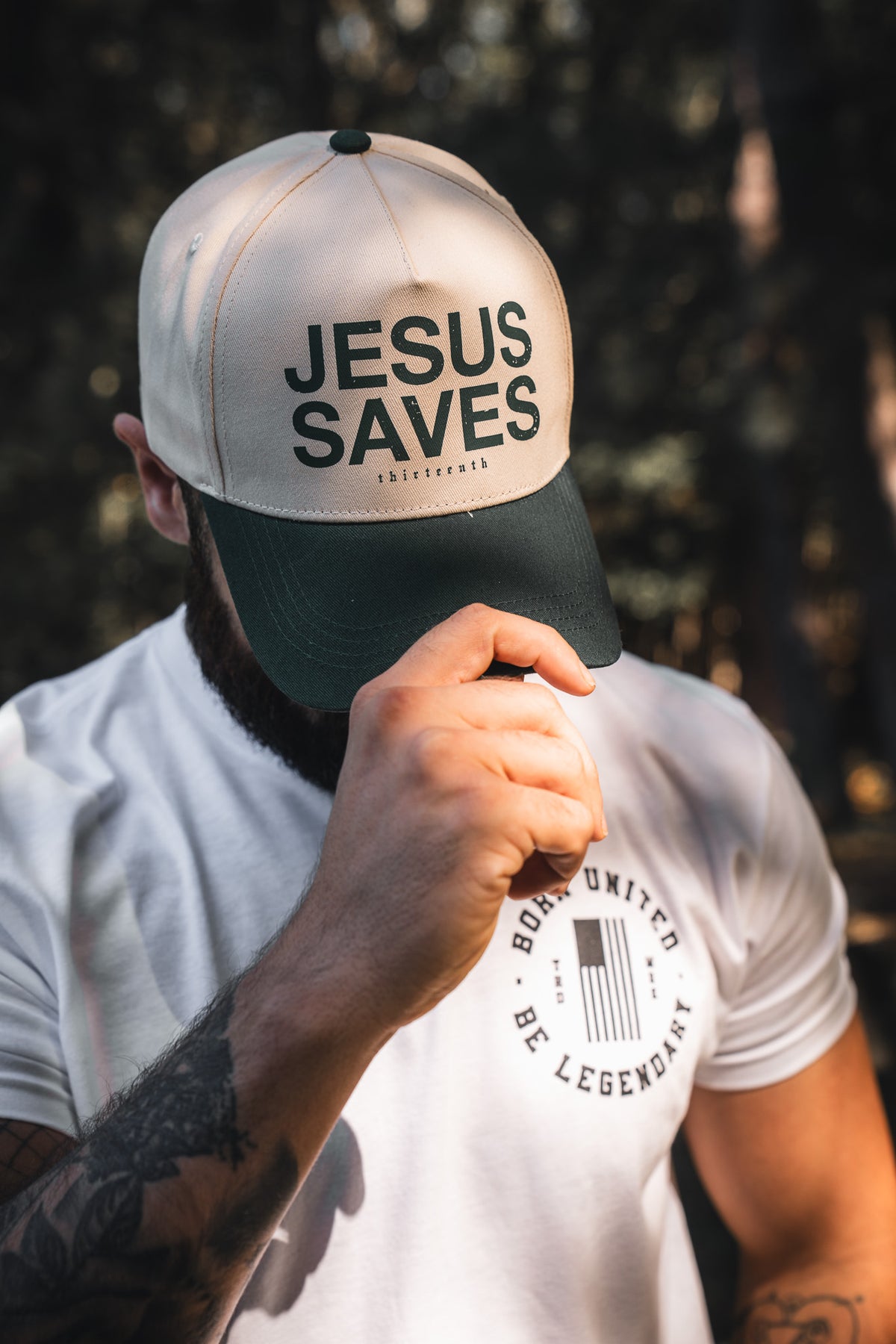 A person adjusts the brim of the 13th Jesus Saves Ivory Snapback outdoors, their face partially hidden by the cap—embroidered with "JESUS SAVES" and referencing John 3:16—and their hand.