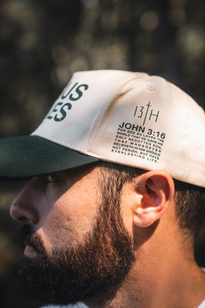 Close-up of a man wearing the 13th Jesus Saves Ivory Snapback, featuring “JOHN 3:16” and a Bible verse above his ear. The softly blurred background is lit with natural light.