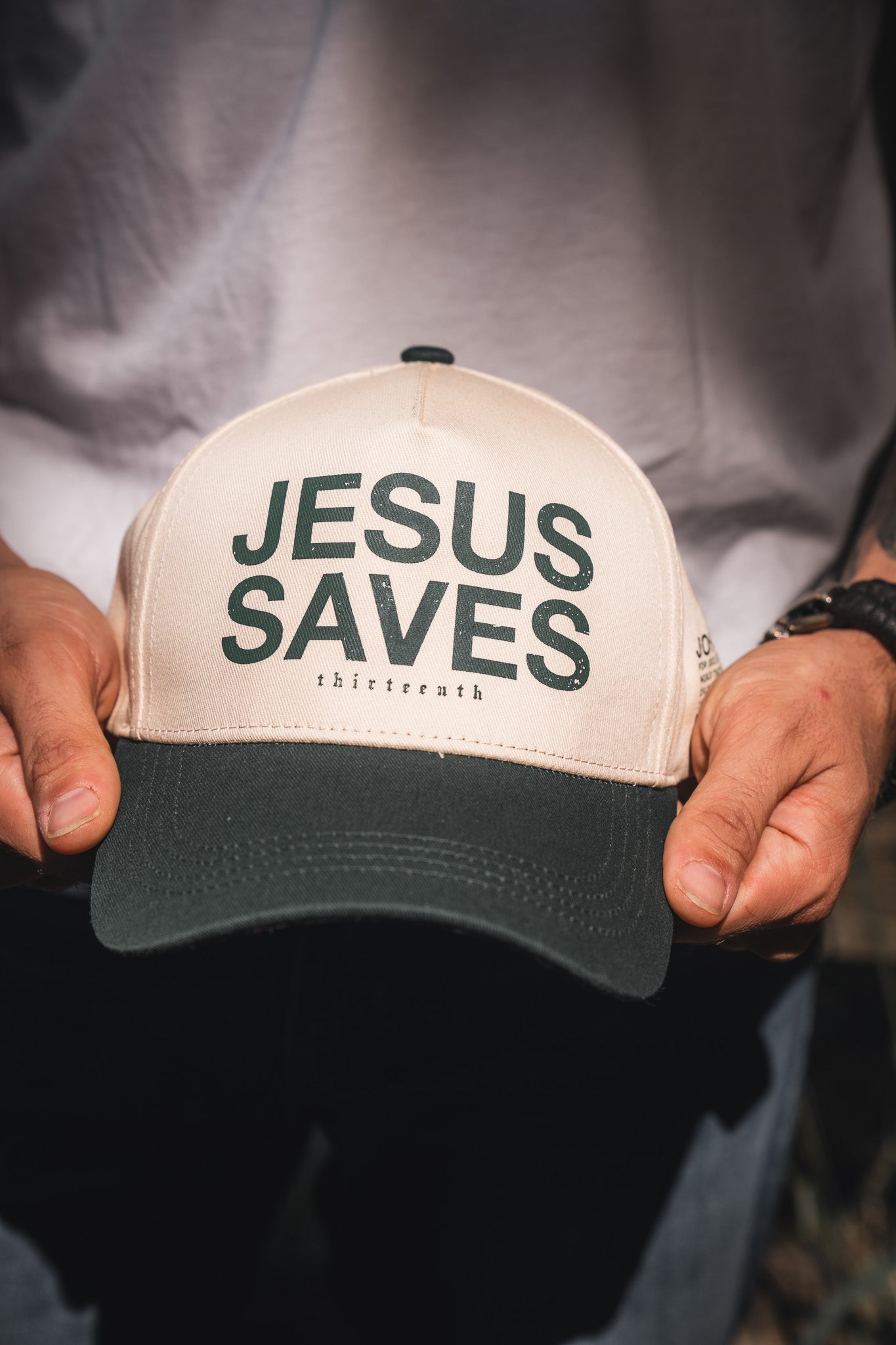 A person wearing a white shirt and watch holds the 13th Jesus Saves Ivory Snapback, a beige and black 5-panel cap with "JESUS SAVES" and "John 3:16" boldly printed on the front.