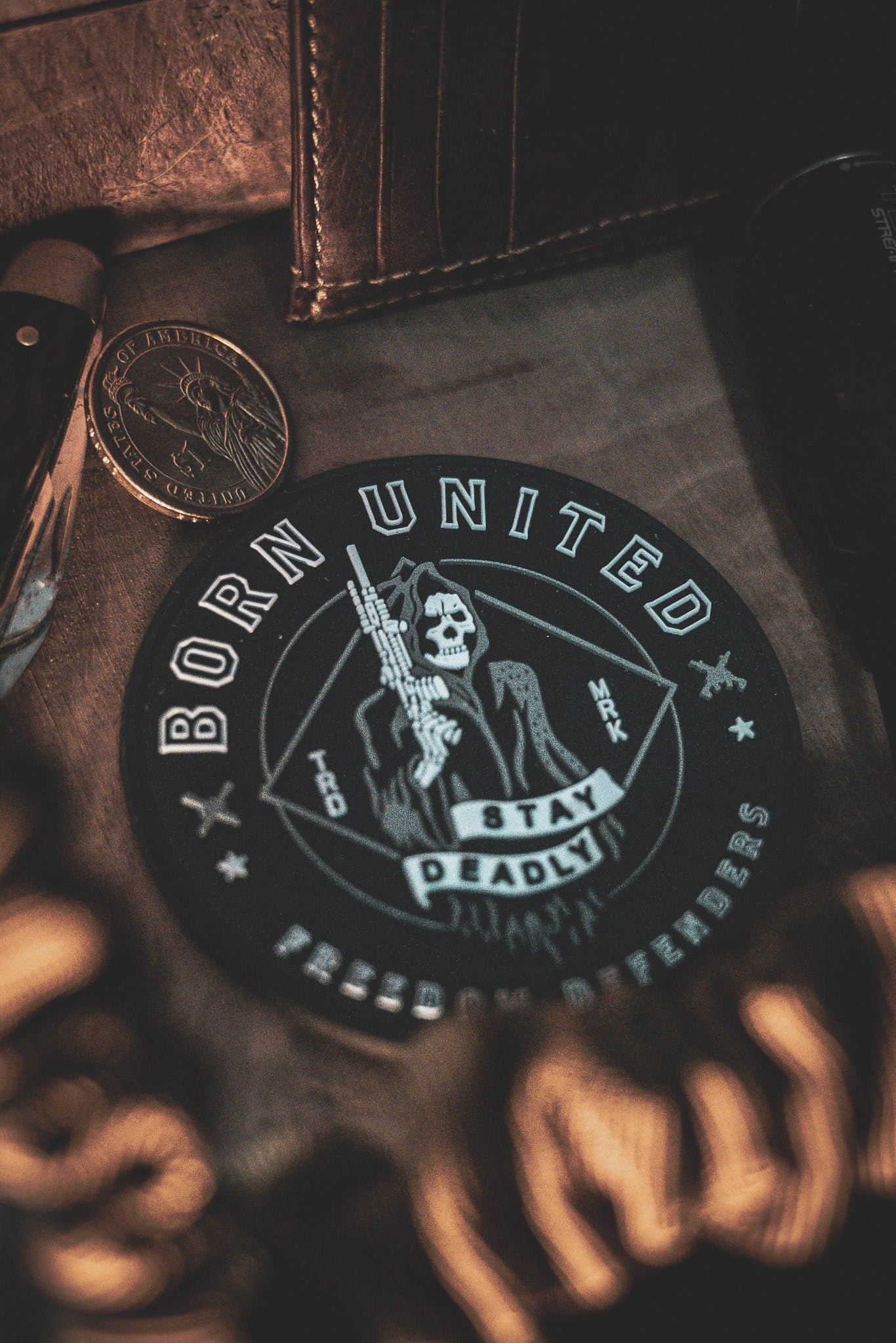 Close-up of the Stay Deadly PVC Patch by Born United Private Label, showcasing a skull with rifle and Born United, Stay Deadly, Freedom Defenders text. Surrounding items include a knife, coin, and wallet on a textured surface under dim dramatic lighting.