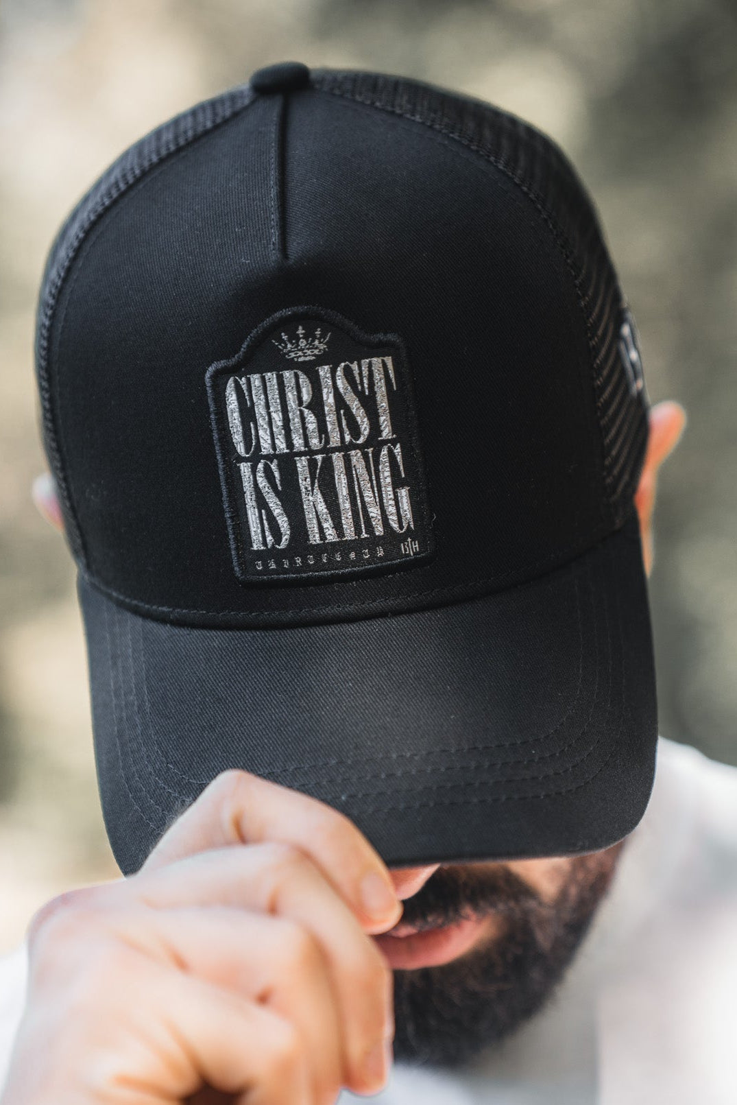A bearded person in a white shirt lowers the brim of a black mesh 13th Christ is King Trucker Snapback, partially obscuring their face and showcasing this stylish handmade hat.
