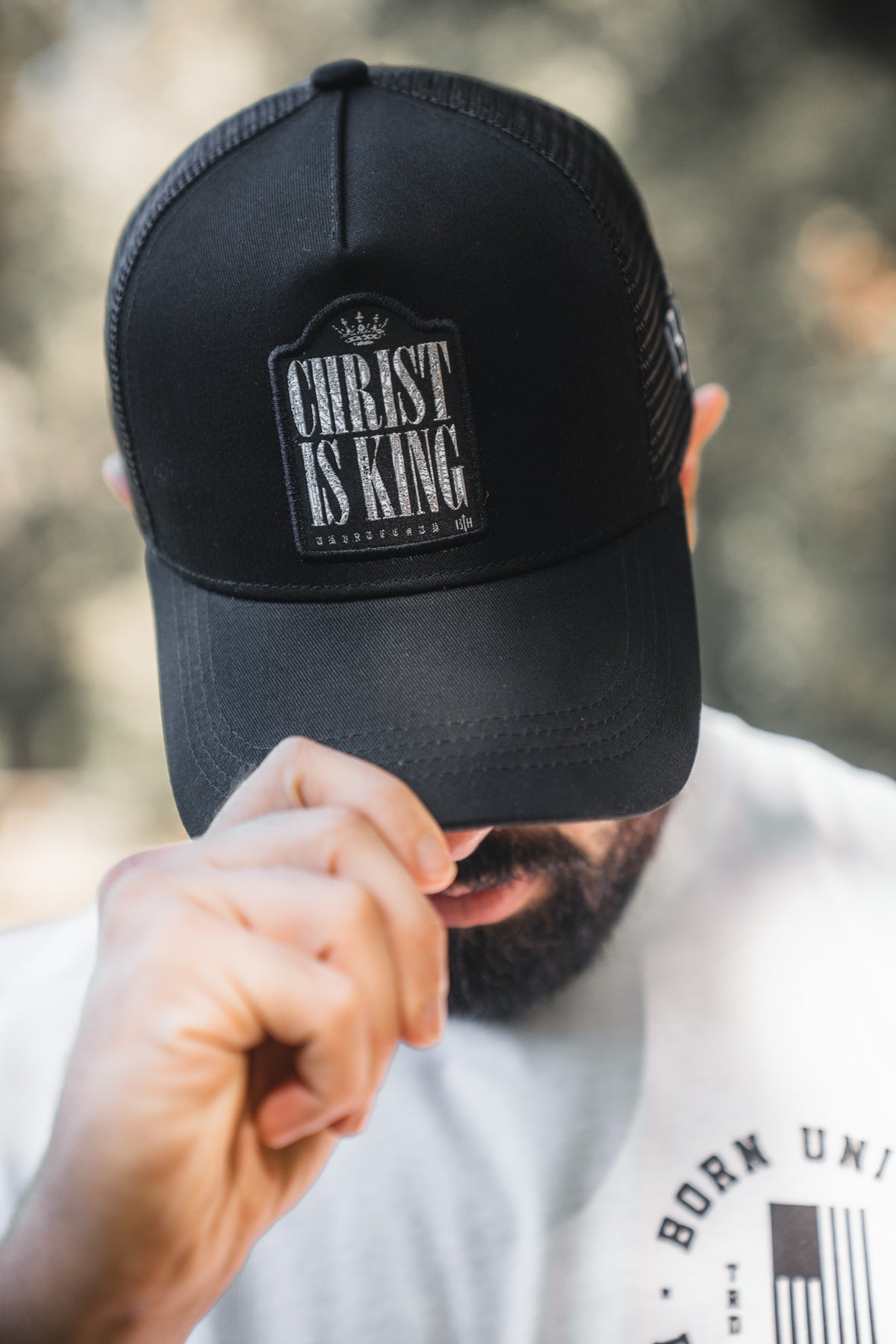 A bearded person in a white shirt lowers the brim of a black mesh 13th Christ is King Trucker Snapback, partially obscuring their face and showcasing this stylish handmade hat.
