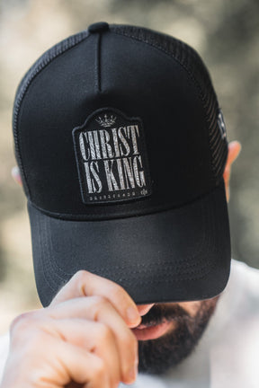 A bearded person in a white shirt lowers the brim of a black mesh 13th Christ is King Trucker Snapback, partially obscuring their face and showcasing this stylish handmade hat.