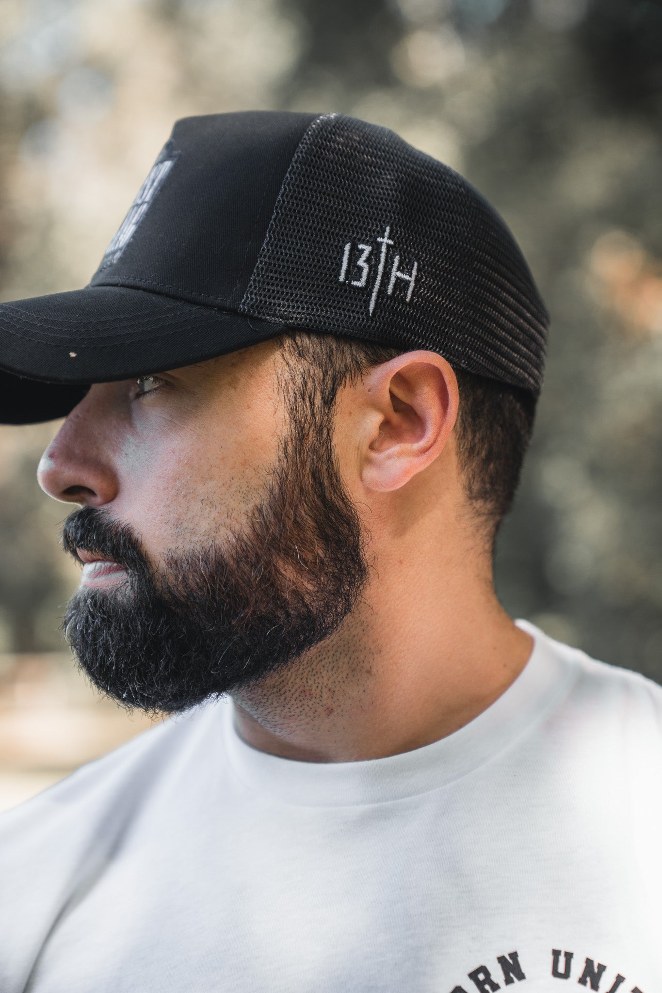 A man with a full beard and mustache, wearing a white shirt and the 13th brand "Christ is King Trucker Snapback" featuring 13TH, a cross, and "Christ is King" embroidery on the side, looks left against a blurred greenery background.