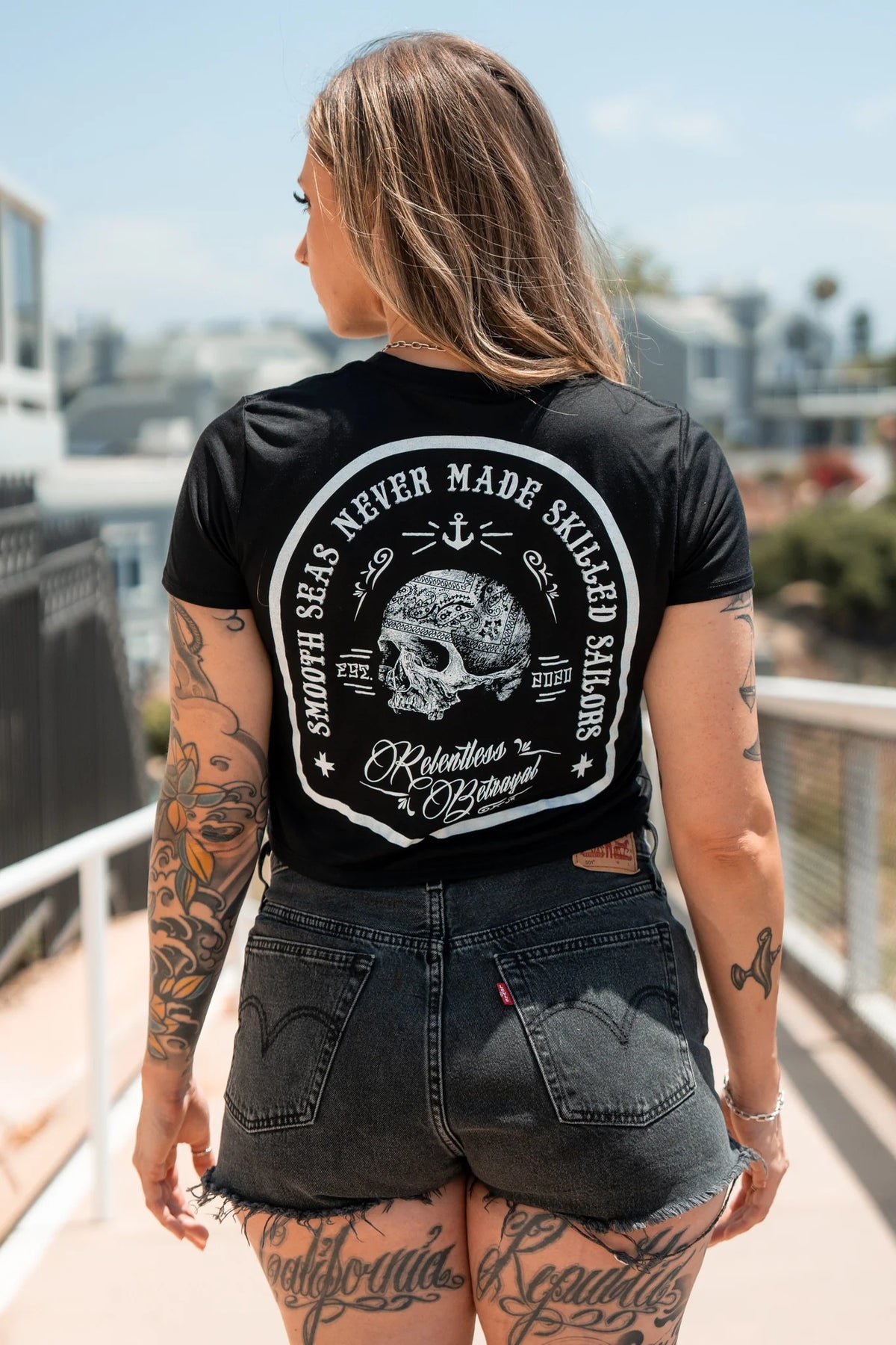 A tattooed individual walking outdoors embraces the pirate lifestyle in a Skilled Sailor Black Crop Tee by Relentless Betrayal, featuring Smooth seas never made skilled sailors with a skull and anchor design. Buildings and trees provide the perfect backdrop.