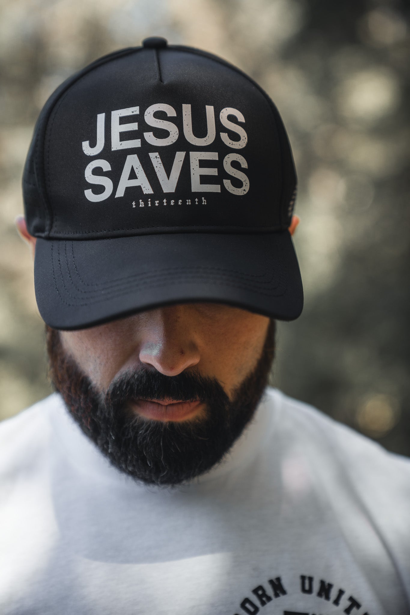 A man with a beard wears a light shirt and the 13th Jesus Saves Black Snapback, boldly embroidered with “JESUS SAVES” in white. He stands outdoors with his head slightly bowed, the softly blurred background enhancing the scene.