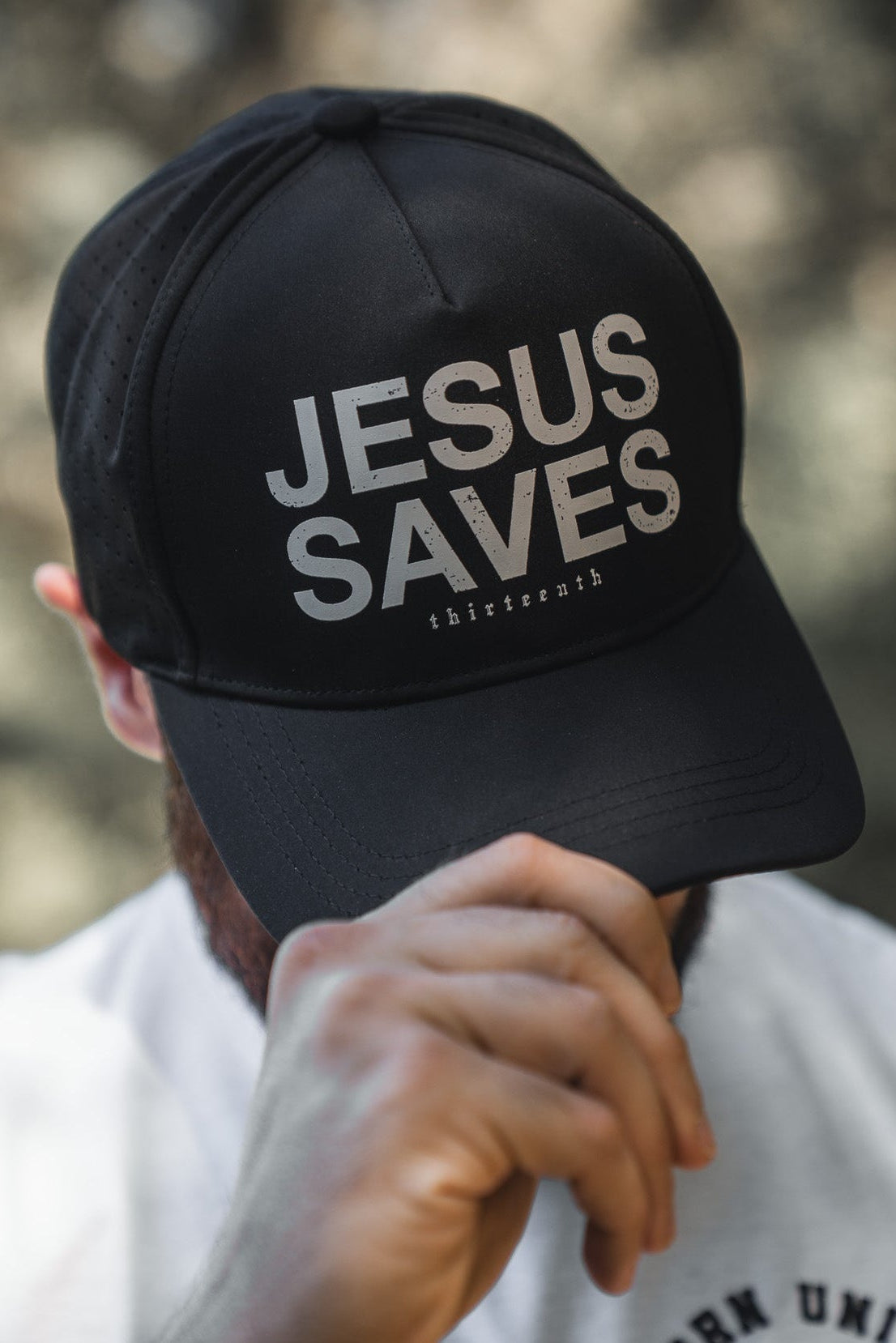A person tilts down their black 13th Jesus Saves Black Snapback to partially cover their face, while wearing a white shirt with "BORN UNITED BE LEGENDARY" printed on it.
