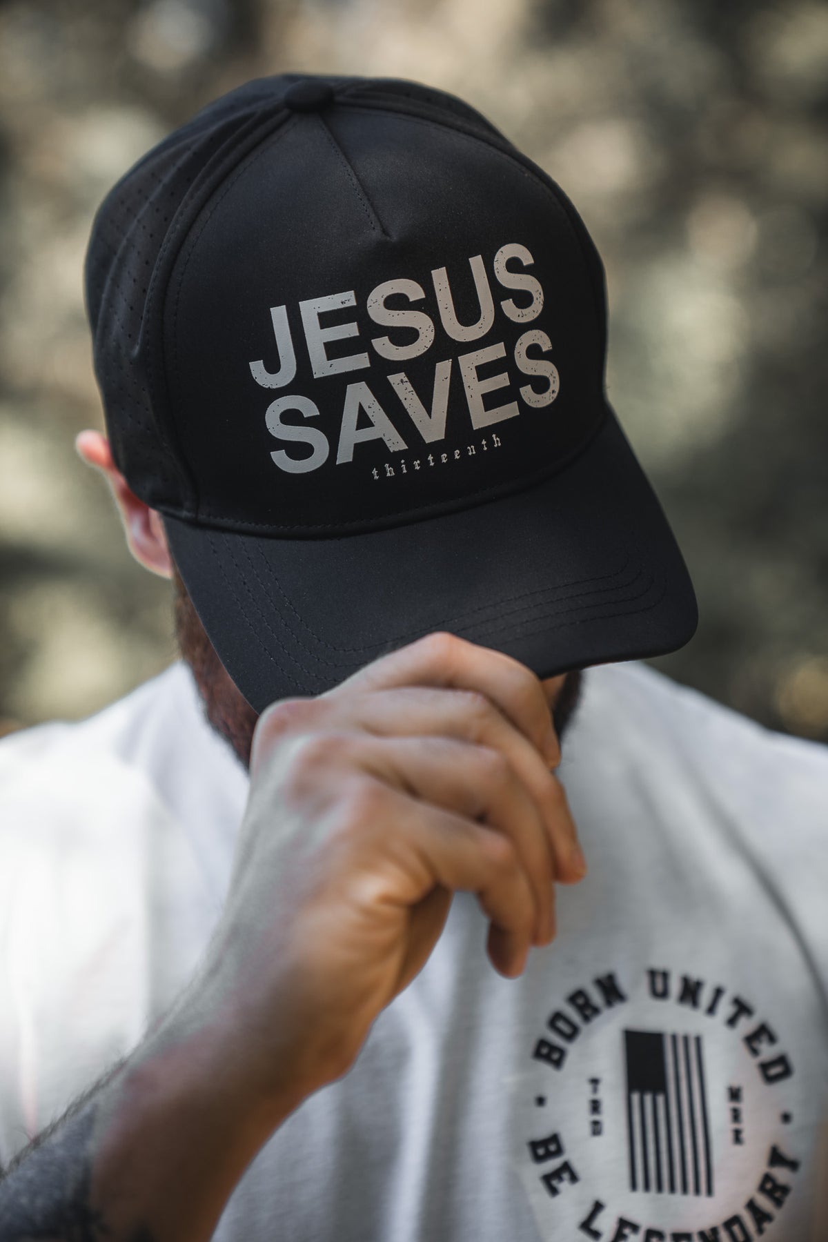 A person tilts down their black 13th Jesus Saves Black Snapback to partially cover their face, while wearing a white shirt with "BORN UNITED BE LEGENDARY" printed on it.