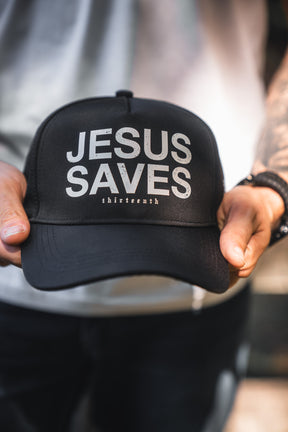 A person holds the Jesus Saves Black Snapback by 13th, featuring bold white "JESUS SAVES" text and smaller "thirteeneeth" lettering. They're dressed in a light shirt and dark pants.