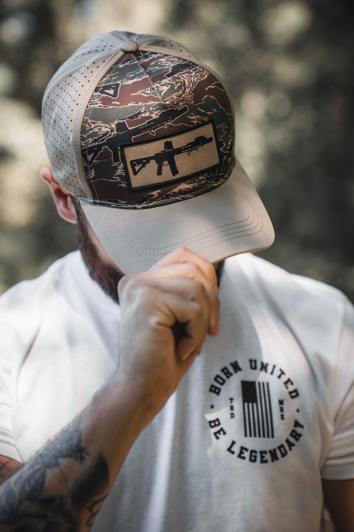 A person outdoors in a camouflage cap with a rifle patch and a white BORN UNITED BE LEGENDARY T-shirt holds the brim—looking ready to share their Legends - Honey I'm Home! treats by Born United Private Label.