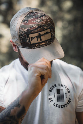 A person outdoors in a camouflage cap with a rifle patch and a white BORN UNITED BE LEGENDARY T-shirt holds the brim—looking ready to share their Legends - Honey I'm Home! treats by Born United Private Label.