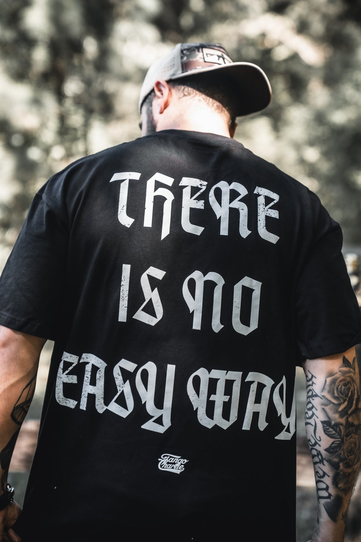 A man stands outdoors with his back to the camera, showing tattooed arms and a cap, wearing a black Tango Charlie Apparel No Easy Way Premium Oversized Tee featuring bold white gothic lettering: THERE IS NO EASY WAY.