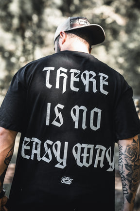 A man stands outdoors with his back to the camera, showing tattooed arms and a cap, wearing a black Tango Charlie Apparel No Easy Way Premium Oversized Tee featuring bold white gothic lettering: THERE IS NO EASY WAY.