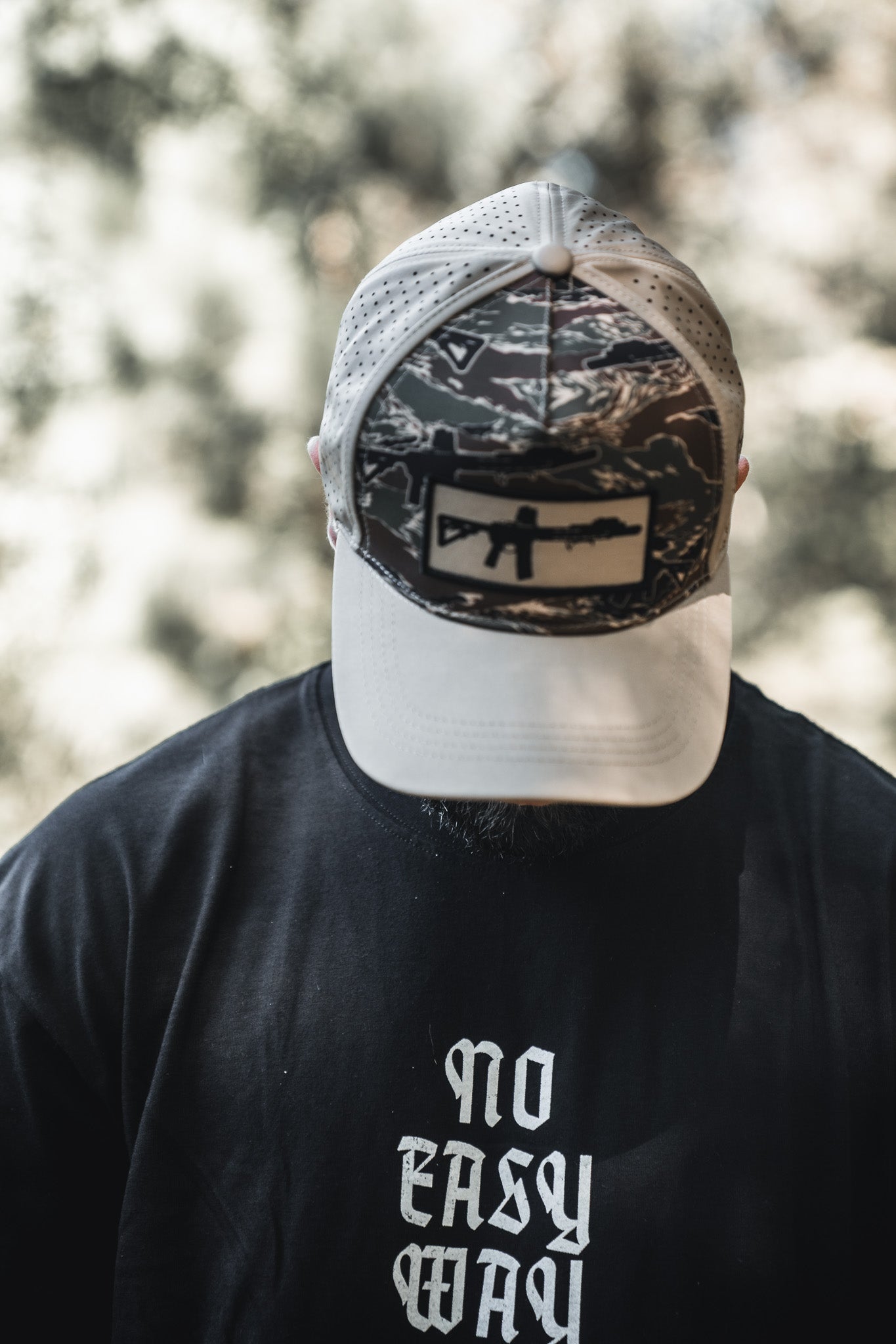 Wearing a Tango Charlie Apparel No Easy Way Premium Oversized Tee and a camouflage cap with rifle graphic, a person stands outdoors among blurred trees, their face mostly hidden by the cap.
