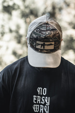 Wearing a Tango Charlie Apparel No Easy Way Premium Oversized Tee and a camouflage cap with rifle graphic, a person stands outdoors among blurred trees, their face mostly hidden by the cap.