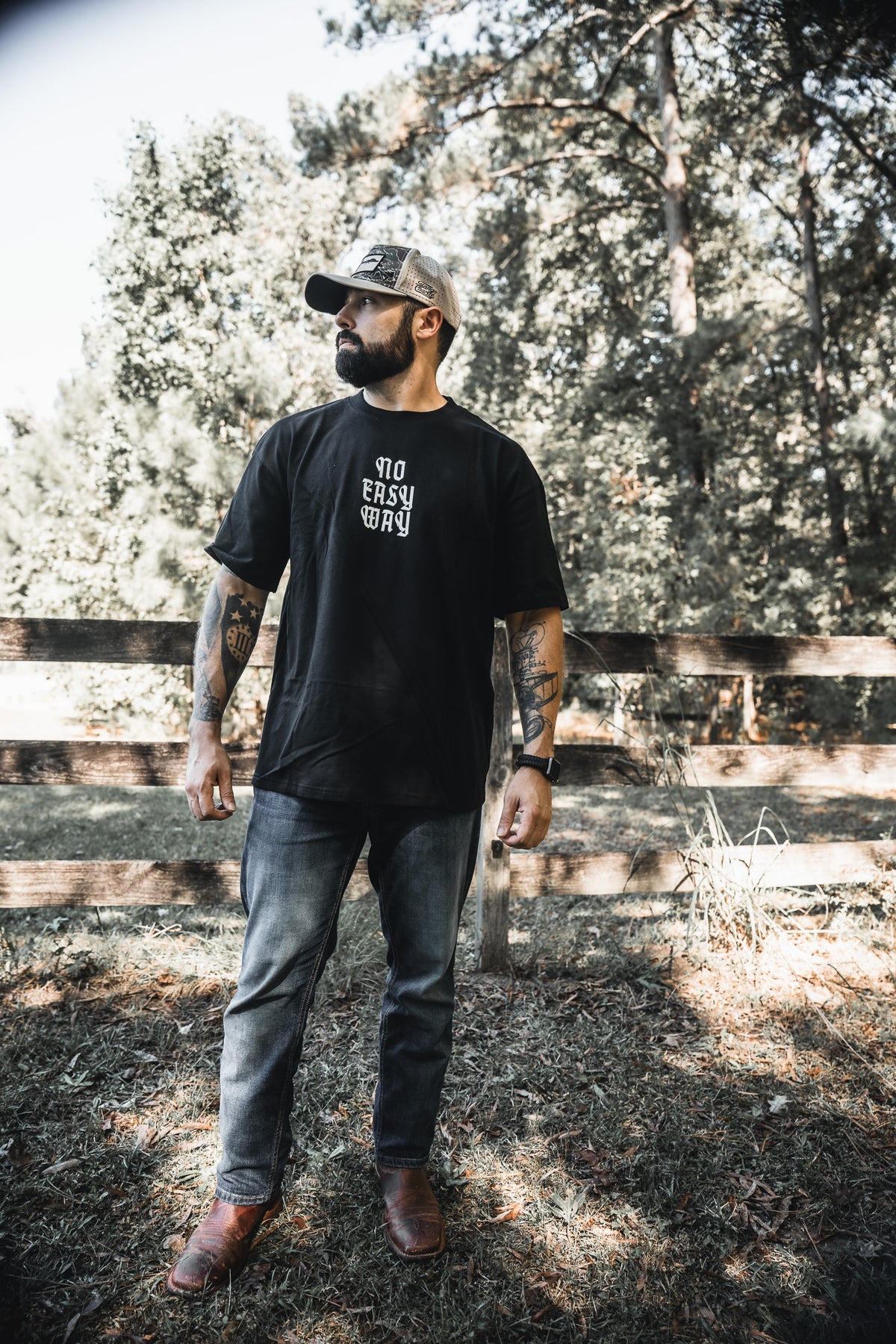 A man with tattoos wears the No Easy Way Premium Oversized Tee by Tango Charlie Apparel, paired with jeans, brown boots, and a baseball cap as he stands on grass near a wooden fence and trees, looking to his left.
