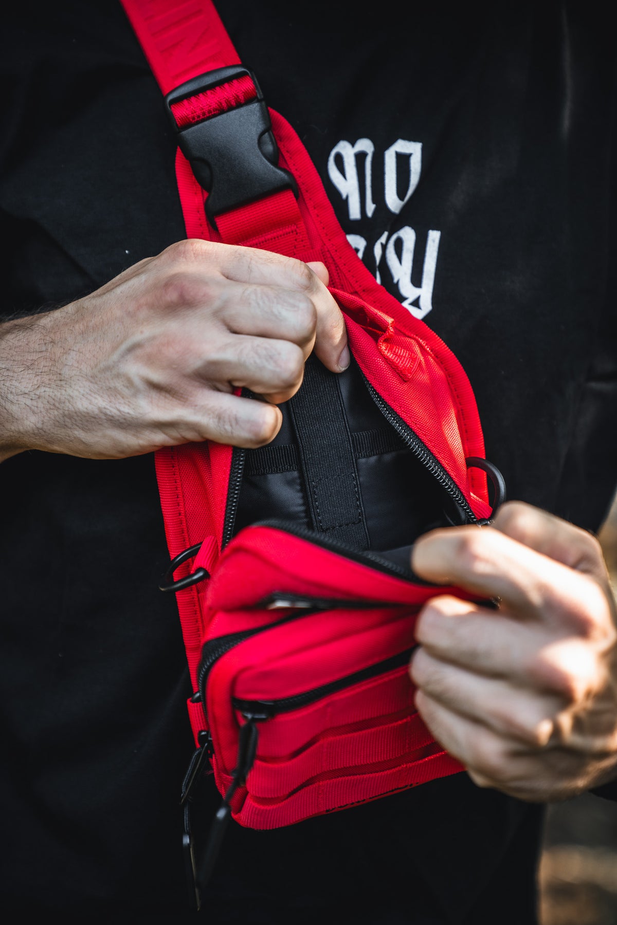 A person in a black shirt opens the zipper of a Born United Private Label Red Reaper Tactical Sling-Pac - 4L, revealing a black item inside. Only the hands, bag, and part of the shirt are visible.