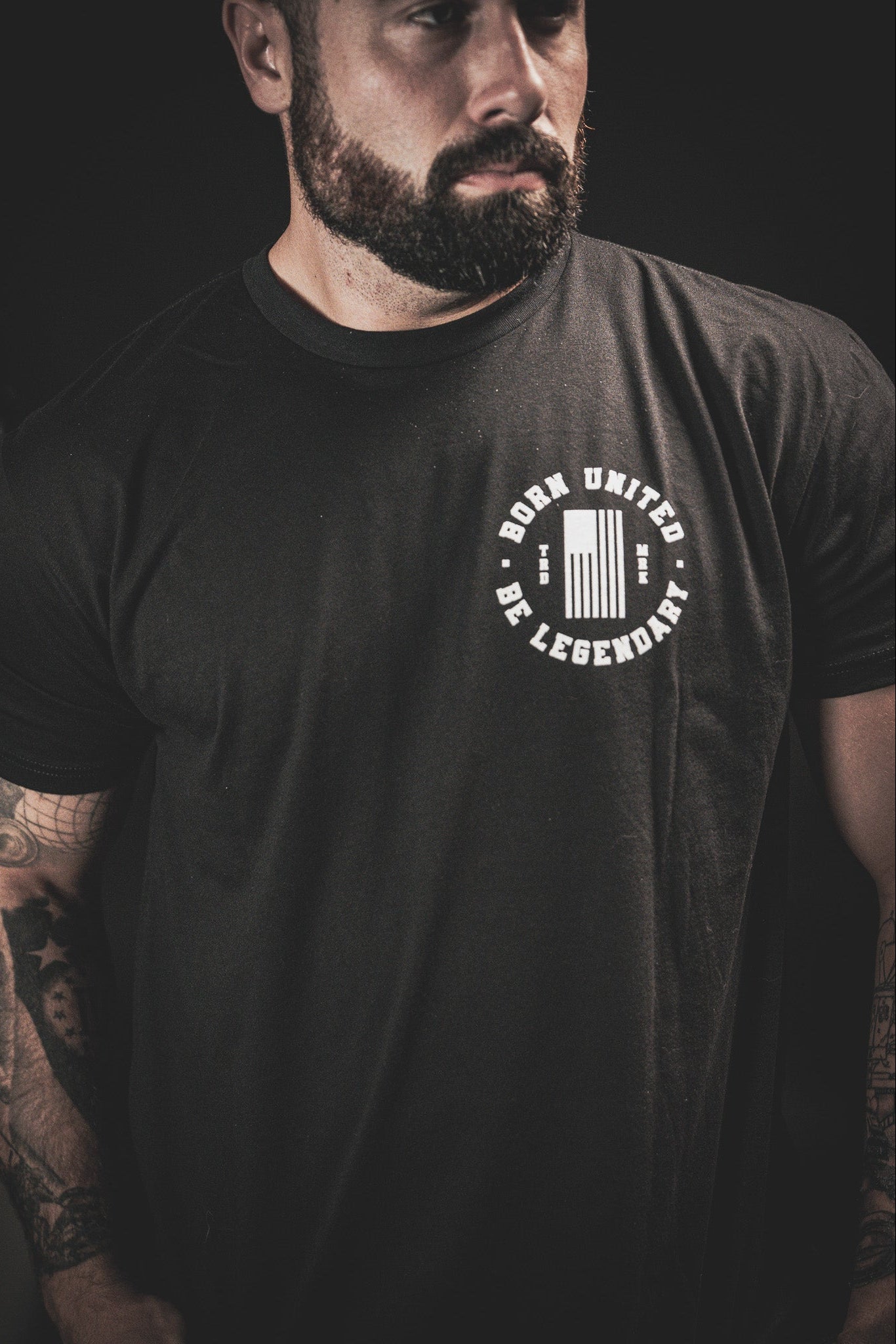 A man with a beard and tattoos on his arms is wearing a black T-shirt with the text Born United Be Legendary and a graphic design. The background is dark and the image is focused on the upper half of his body.