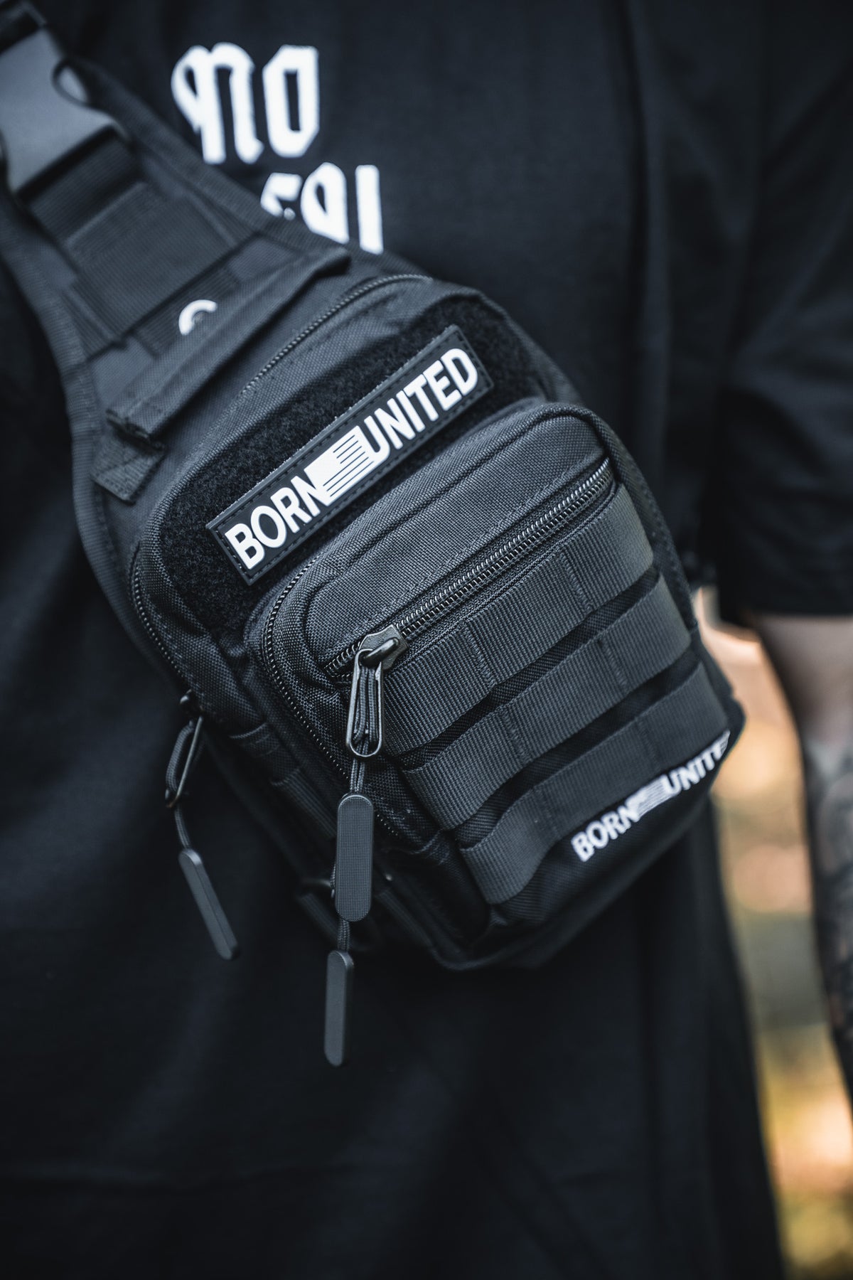 Close-up of the Born United Private Label Black Phantom Tactical Sling-Pac - 4L, featuring several zippers and straps, worn over a black shirt with BORN UNITED patches. The wearer's tattooed arm is partially visible.
