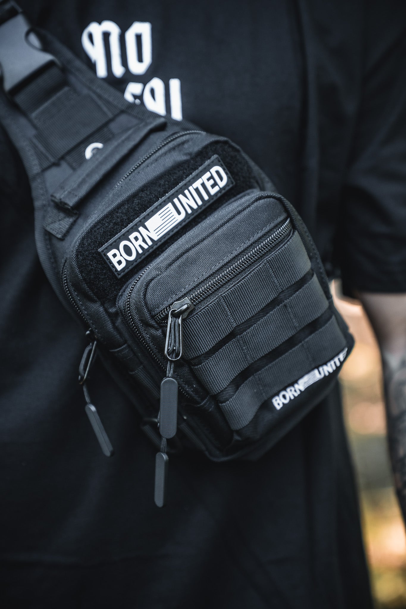 Close-up of the Born United Private Label Black Phantom Tactical Sling-Pac - 4L, featuring several zippers and straps, worn over a black shirt with BORN UNITED patches. The wearer's tattooed arm is partially visible.