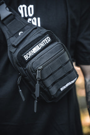 Close-up of the Born United Private Label Black Phantom Tactical Sling-Pac - 4L, featuring several zippers and straps, worn over a black shirt with BORN UNITED patches. The wearer's tattooed arm is partially visible.