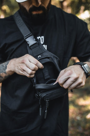 A man in a black t-shirt and watch opens the zipper of a Born United Private Label Black Phantom Tactical Sling-Pac - 4L outdoors, revealing its contents; his face is partially hidden by a hat.