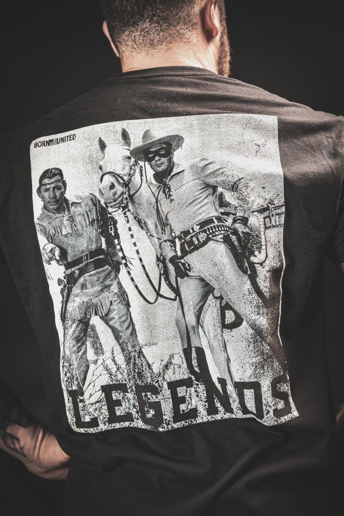 Back of a person wearing a black T-shirt featuring a vintage-style print of two cowboys from an old Western, alongside a horse. The word LEGENDS is printed at the bottom, and the word BORNLIMITED is above the image.