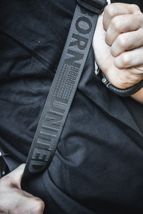 A close-up shows a person in a black shirt fastening a seatbelt marked "UNITED" and sporting the stylized flag. They carry the Born United Private Label Black Phantom Tactical Sling-Pac - 4L for easy concealed carry and daily essentials.