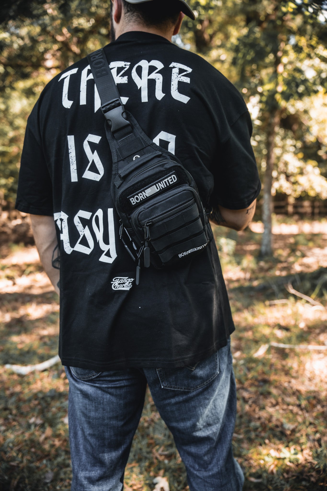 Outdoors, a person wears jeans, a black shirt with bold white back text, and the Born United Private Label Black Phantom Tactical Sling-Pac - 4L. Trees and sunlight are in the background.
