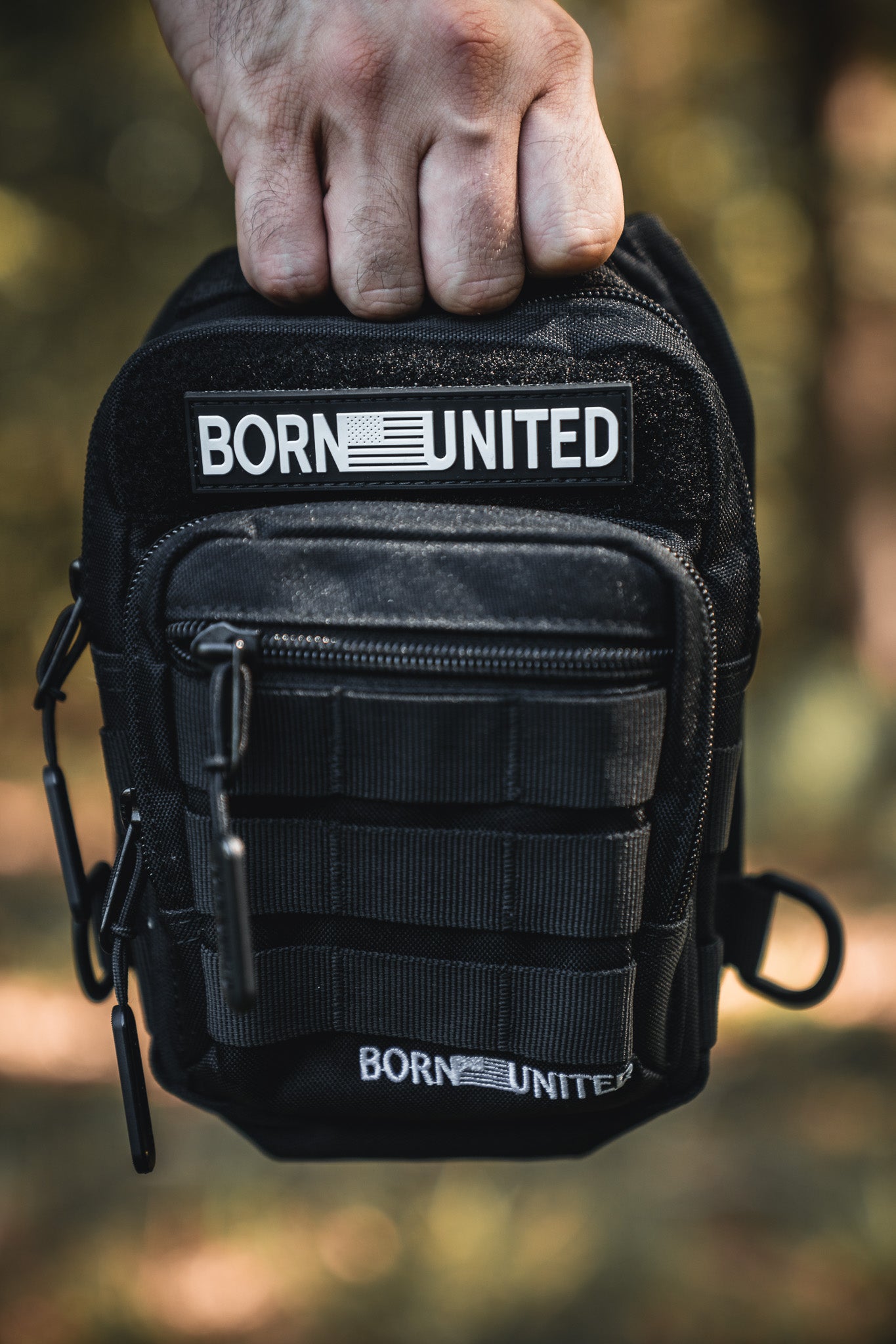A hand holds the Born United Private Label Black Phantom Tactical Sling-Pac - 4L, featuring zippers, MOLLE webbing, and two BORN UNITED patches—one at the top and one at the bottom—against a blurred outdoor backdrop.