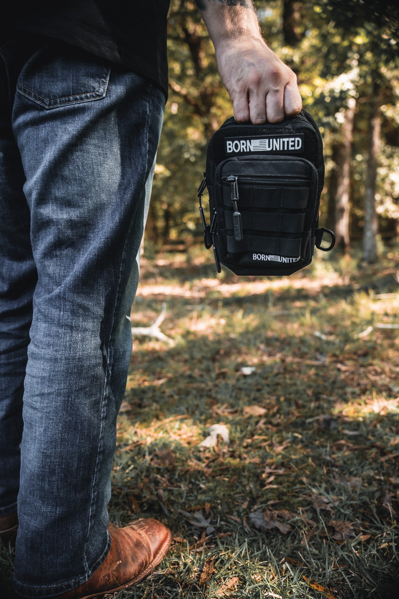 In a wooded area, only the lower body and hand of a person are visible as they hold the Black Phantom Tactical Sling-Pac - 4L by Born United Private Label. They stand on grass, wearing blue jeans and brown shoes.