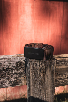 The BU Logo Belt - Brown by Born United Private Label, featuring a black buckle, is displayed on a weathered wooden fence post with sunlight illuminating a reddish wooden wall in the background.