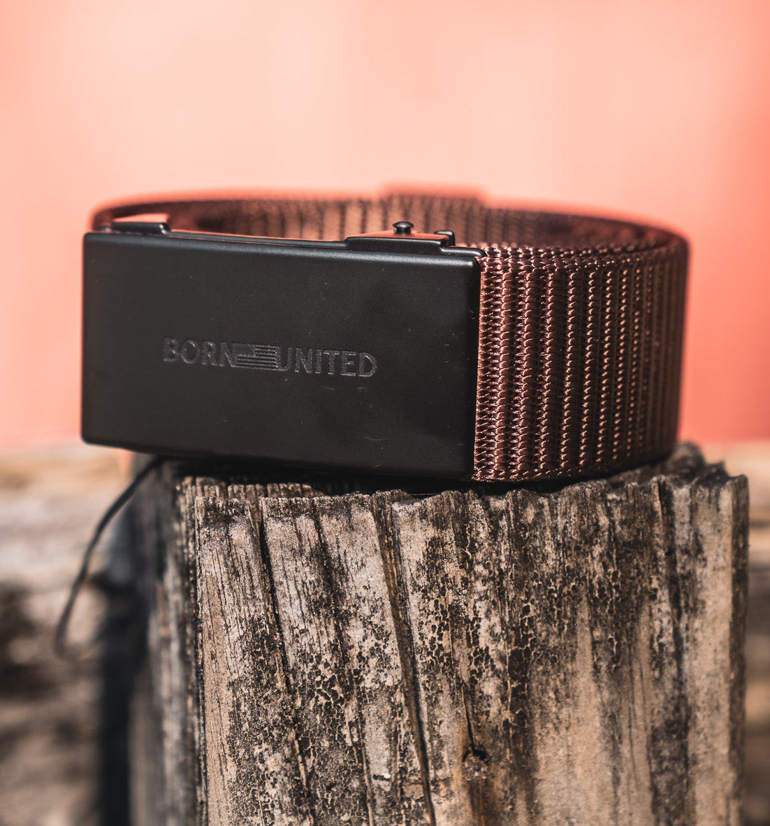 The BU Logo Belt - Brown by Born United Private Label features a brown woven design, black engraved buckle, and adjustable fit. Coiled on a rustic wooden post with a warm blurred background, it blends style with comfort.