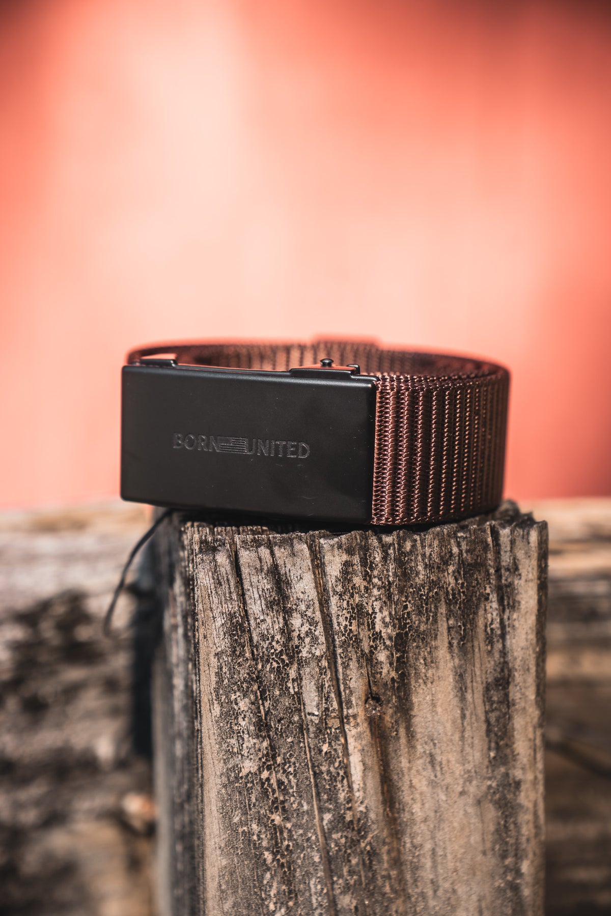 The BU Logo Belt - Brown by Born United Private Label features a brown woven design, black engraved buckle, and adjustable fit. Coiled on a rustic wooden post with a warm blurred background, it blends style with comfort.