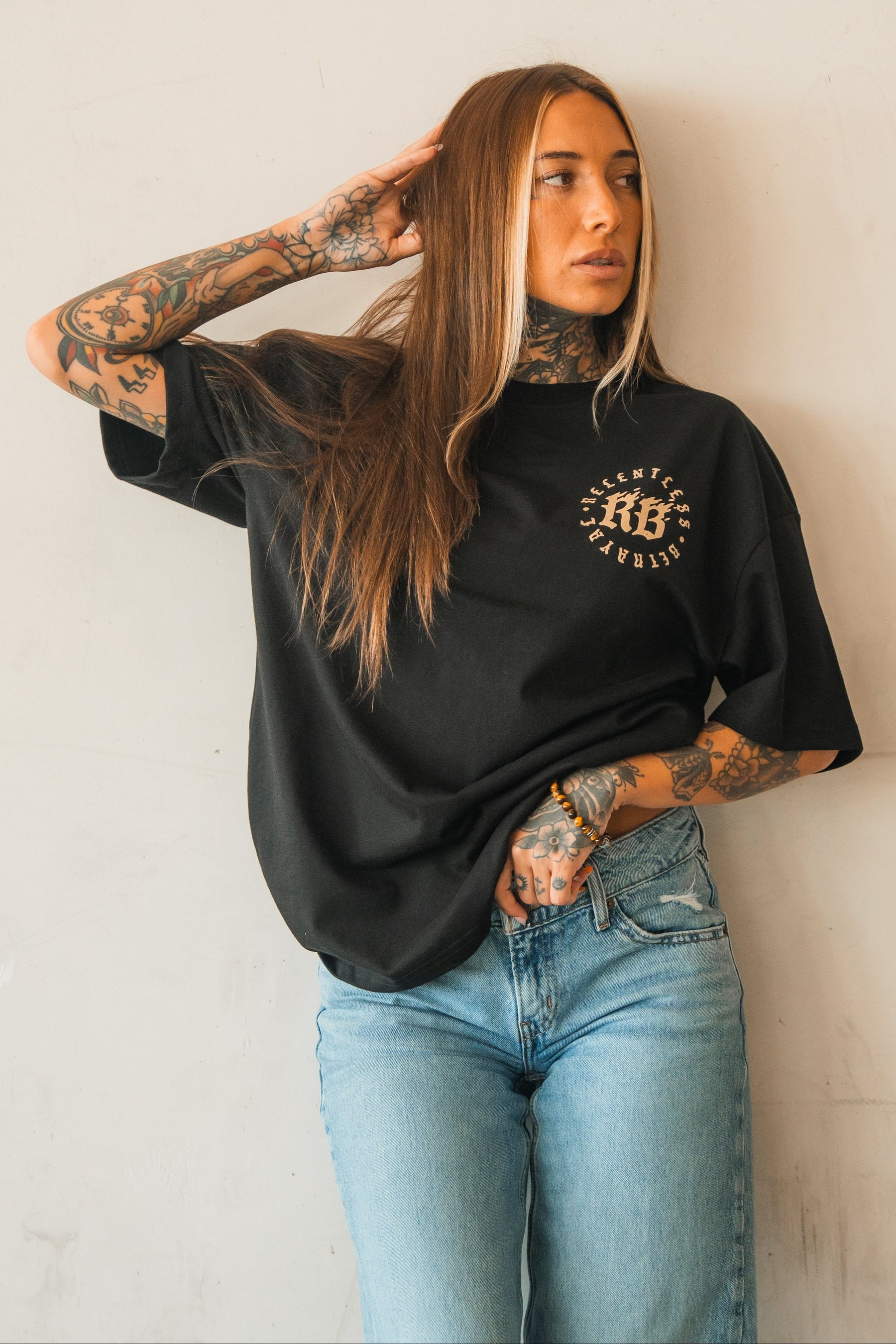 A woman with long brown hair and tattoos poses against a light wall, wearing the Relentless Betrayal Never Surrender Premium Oversized Ghost T-Shirt and light blue jeans. She rests one hand on her head and gazes to the side.